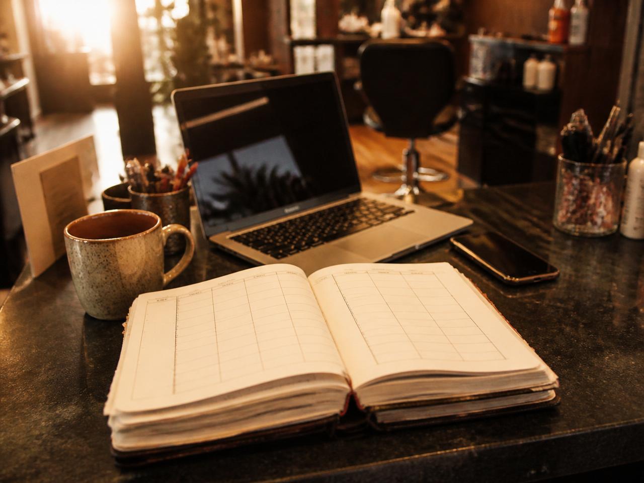 Paper appointment book open on a salon front desk beside a laptop and a phone in warm morning light